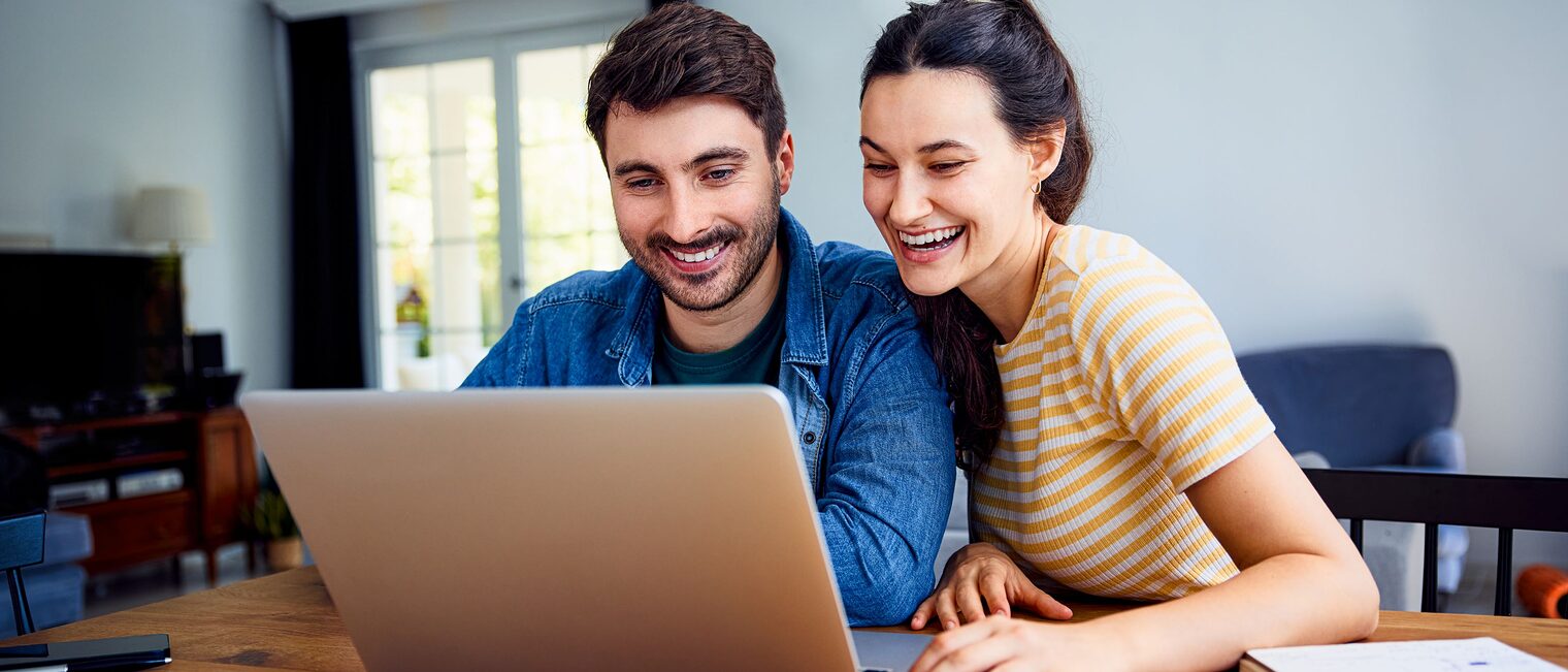 Frau und Mann sitzen an einem Tisch und blicken in einen Laptop. Junge Frau und junger Mann sitzen zusammen an einem Tisch und blicken mit Freude in einen Laptop.